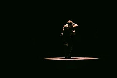 Scène uit HABITAT: een danser staat gebogen in een enkele lichtcirkel op een verder donker podium, met zijn armen om zijn hoofd geslagen.