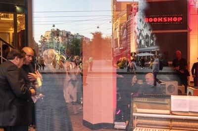 Beeld bij Electronic Music and the Present: uitzicht door een grote ruit op een plein bij avondlicht, met rechts een rode lichtreclame met het woord 'BOOKSHOP'. In het glas weerspiegelen bezoekers met een drankje in de hand.
