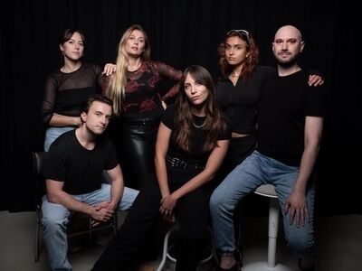Groepsportret van zes spelers van XTC: the Electronical Musical in zwarte kleding tegen een donkere achtergrond, deels zittend op krukken.