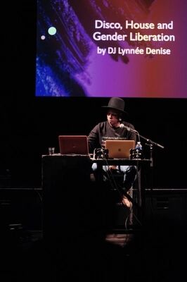 Beeld bij Rooted in Culture: UK Sounds influencing House culture: een dj met een brede zwarte hoed zit achter een tafel met twee laptops en dj-controllers, met een microfoon voor zich. Op het scherm erboven staat 'Disco, House and Gender Liberation by DJ Lynnée Denise'.